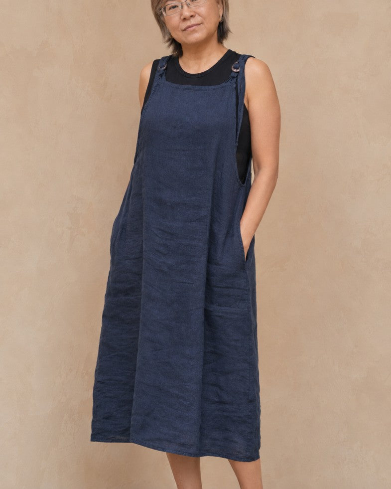 A woman wearing a sleeveless navy blue linen pinafore dress over a black crewneck top, standing against a textured beige wall.