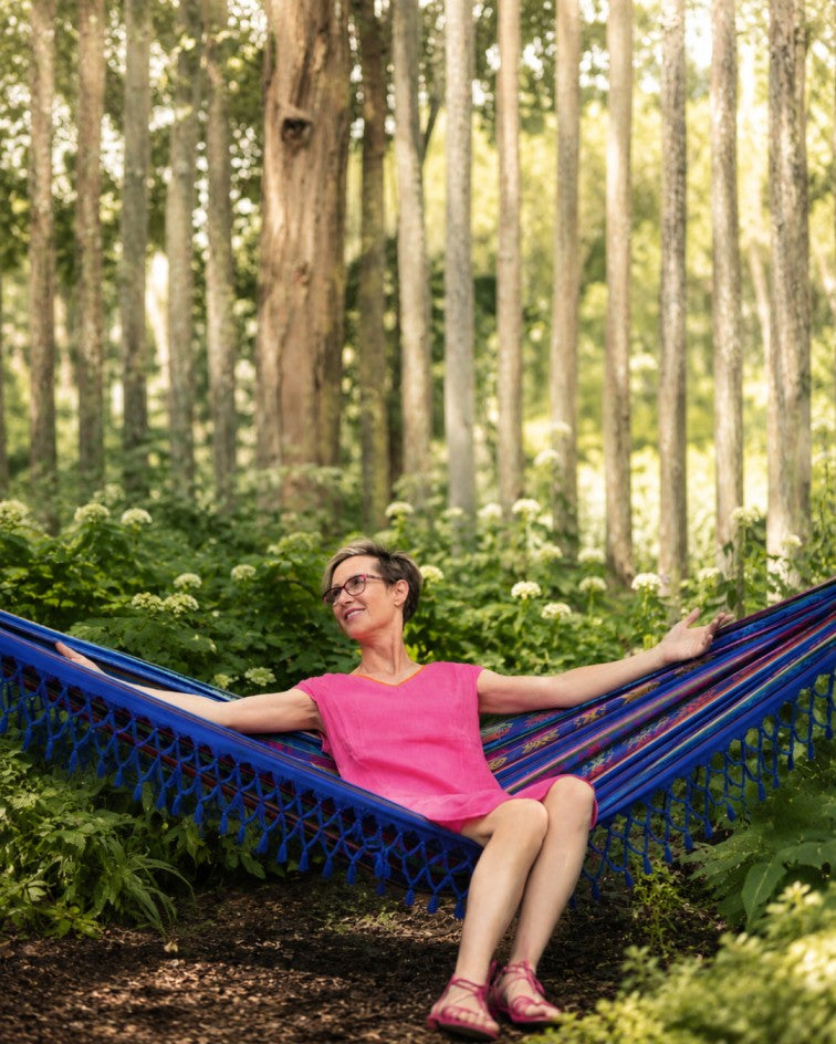 A woman wearing a bright pink dress sits comfortably in a blue patterned hammock strung between trees in a lush green forest.
