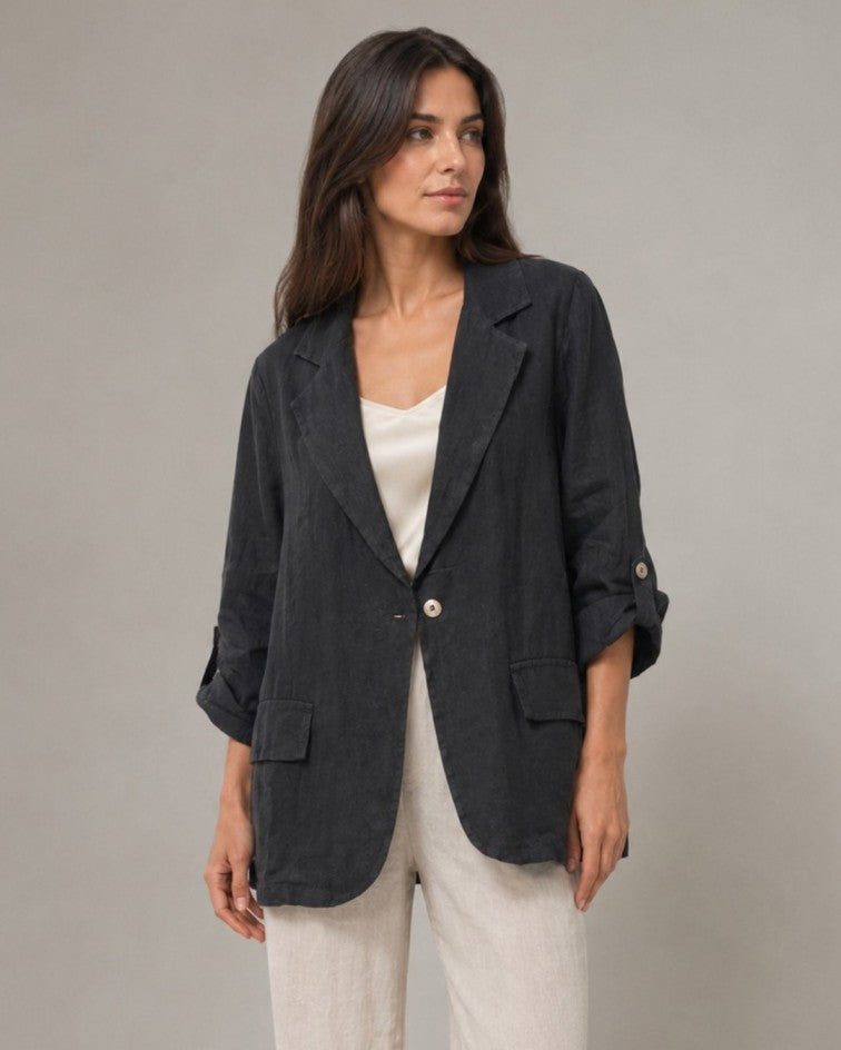 A woman wearing a dark charcoal gray linen-blend blazer over a white top and light beige trousers, standing against a neutral gray studio background.